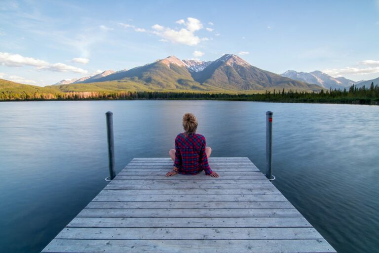 Femme assise au bout d’un ponton sur un lac, contemplative face aux montagnes ensoleillées.