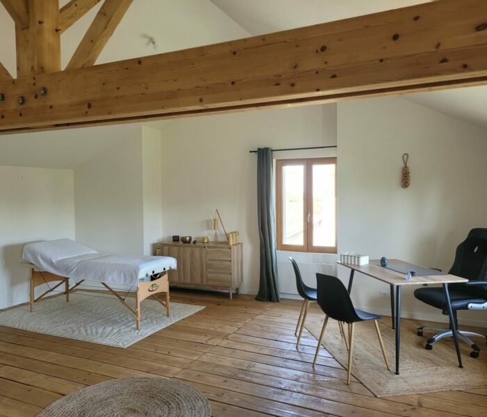 Espace de bien-être à Chemillé avec poutre apparente, parquet, table de massage sur un tapis, meuble avec bol chantant et carillon Koshi, fenêtre et bureau
